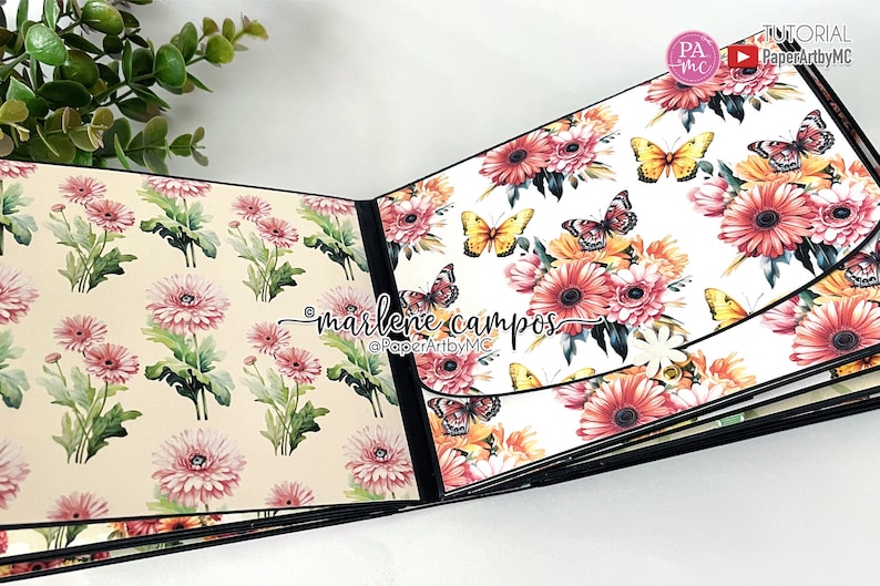 May include: A scrapbook with a floral design featuring pink and white flowers and butterflies. The scrapbook is open to reveal a page with a white background and a floral design featuring pink, yellow, and orange flowers and butterflies. The text "marlene campos" and "@PaperArtbyMC" is printed on the page.