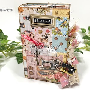 May include: A handmade sewing journal with a patchwork design. The cover features a metal label that reads "SEWING". Embellishments include fabric scraps, lace, and a miniature sewing machine charm. The journal is decorated with images of sewing machines, scissors, and roses.