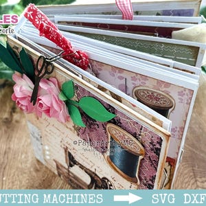 Op de afbeelding: Een meerbladig plakboekalbum met een naaithema, voorzien van illustraties van garenklosjes en een naaimachine. De kaft is versierd met roze papieren bloemen, groene bladeren, een rood stoffen lint en een kleine metalen schaarbedel.