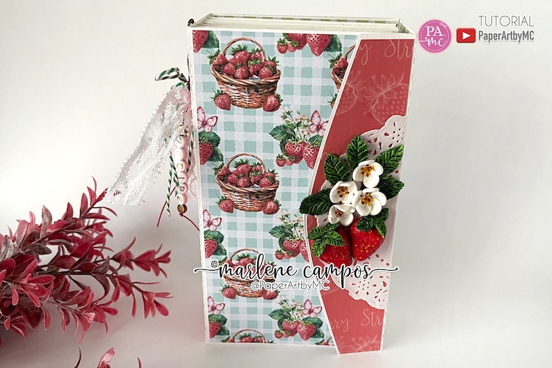 May include: A handmade journal with a strawberry theme. The cover features a basket of strawberries on a light blue gingham pattern. The journal is decorated with paper strawberries, white flowers, and lace. The text "marlene campos" is visible.