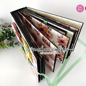 May include: A black and white floral scrapbook with a green ribbon. The scrapbook is closed and has a floral design on the cover. The scrapbook is decorated with flowers and butterflies.