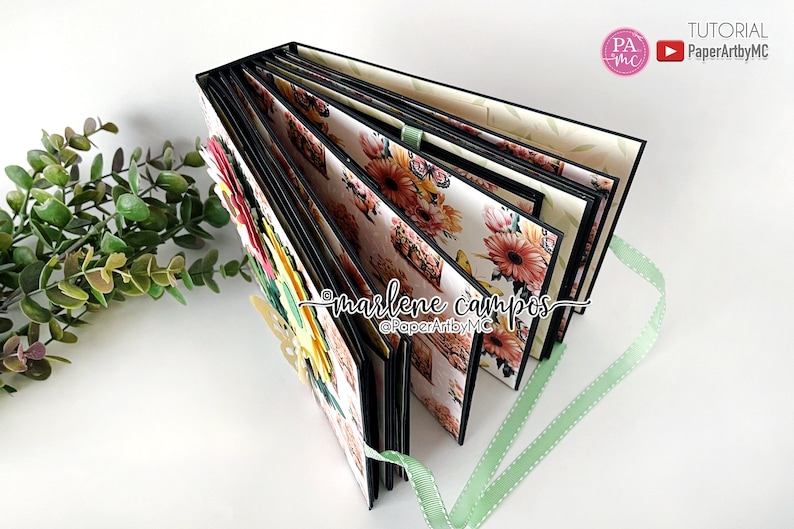May include: A black and white floral scrapbook with a green ribbon. The scrapbook is closed and has a floral design on the cover. The scrapbook is decorated with flowers and butterflies.