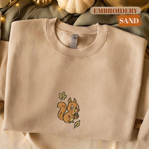 May include: Sand-colored sweatshirt with an embroidered squirrel holding an acorn. The squirrel is brown with a white belly and is surrounded by green leaves. The text "EMBROIDERY SAND" is visible in the background.