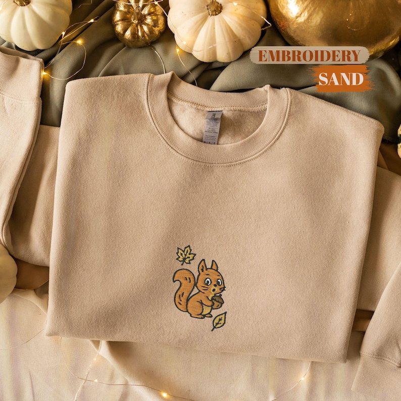 May include: Sand-colored sweatshirt with an embroidered squirrel holding an acorn. The squirrel is brown with a white belly and is surrounded by green leaves. The text "EMBROIDERY SAND" is visible in the background.