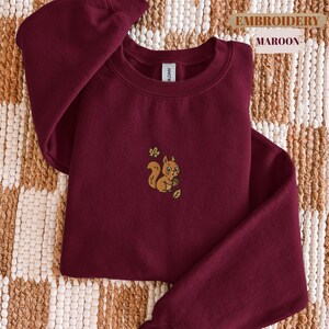 May include: Maroon sweatshirt with an embroidered squirrel design. The squirrel is holding a nut and is surrounded by leaves. The text "EMBROIDERY" and "MAROON" are printed on the top right corner of the image.