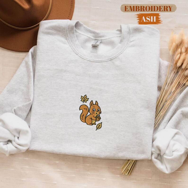 May include: A light gray sweatshirt with a crew neck featuring an embroidered squirrel holding a nut and surrounded by leaves. The text "EMBROIDERY ASH" is in the upper right corner. The sweatshirt is next to a brown hat and dried wheat.