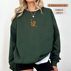 May include: Forest green sweatshirt with a white undershirt. The sweatshirt features an embroidered squirrel design with the word "EMBROIDERY" above the design. The words "FOREST GREEN" are also visible.
