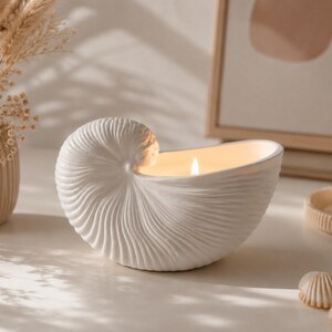 Peut inclure: Un bougeoir en céramique blanche en forme de coquille de nautile avec une surface texturée et nervurée. Le récipient contient une bougie allumée avec une flamme visible, diffusant une lueur chaude depuis l'intérieur de la structure en spirale.