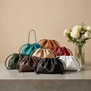 Può includere: Una collezione di borse a coulisse in vari colori, tra cui turchese, marrone, nero, bianco e rosso. Alcune borse presentano tracolle a catena, altre chiusure a coulisse. Un vaso di rose bianche è nelle vicinanze.