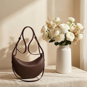 Può includere: Una borsa a mezzaluna marrone scuro con una lunga tracolla regolabile. La borsa ha una cerniera dorata e una texture liscia. Accanto alla borsa c'è un vaso bianco con fiori bianchi.