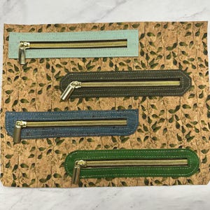 May include: Four rectangular zipper panels in shades of blue, green, and grey, each with a gold-coloured zip. The panels are set against a corkboard background with a leaf pattern.