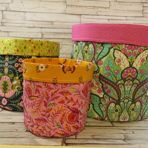 May include: Three fabric storage baskets with colorful floral patterns. The largest basket is pink with green and white floral designs, the middle basket is pink with orange and yellow floral designs, and the smallest basket is black with green and pink floral designs.