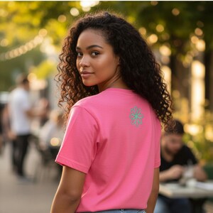 Puede incluir: Una camiseta rosa con un dise&ntilde;o floral turquesa en la espalda. La camiseta de manga corta est&aacute; hecha de un material suave. La persona de la imagen est&aacute; mirando por encima del hombro.