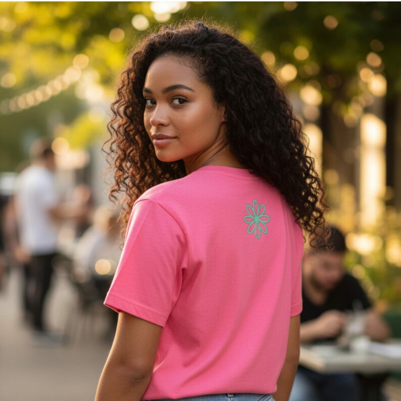 Puede incluir: Una camiseta rosa con un dise&ntilde;o floral turquesa en la espalda. La camiseta de manga corta est&aacute; hecha de un material suave. La persona de la imagen est&aacute; mirando por encima del hombro.