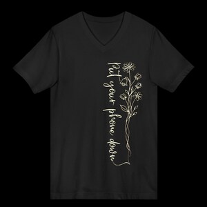 Puede incluir: Camiseta negra con cuello en V con un dise&ntilde;o floral y el texto "Put your phone down" en escritura vertical. El dise&ntilde;o presenta un delicado dibujo lineal de flores y hojas en color oro claro.