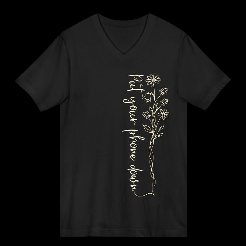 Puede incluir: Camiseta negra con cuello en V con un dise&ntilde;o floral y el texto "Put your phone down" en escritura vertical. El dise&ntilde;o presenta un delicado dibujo lineal de flores y hojas en color oro claro.