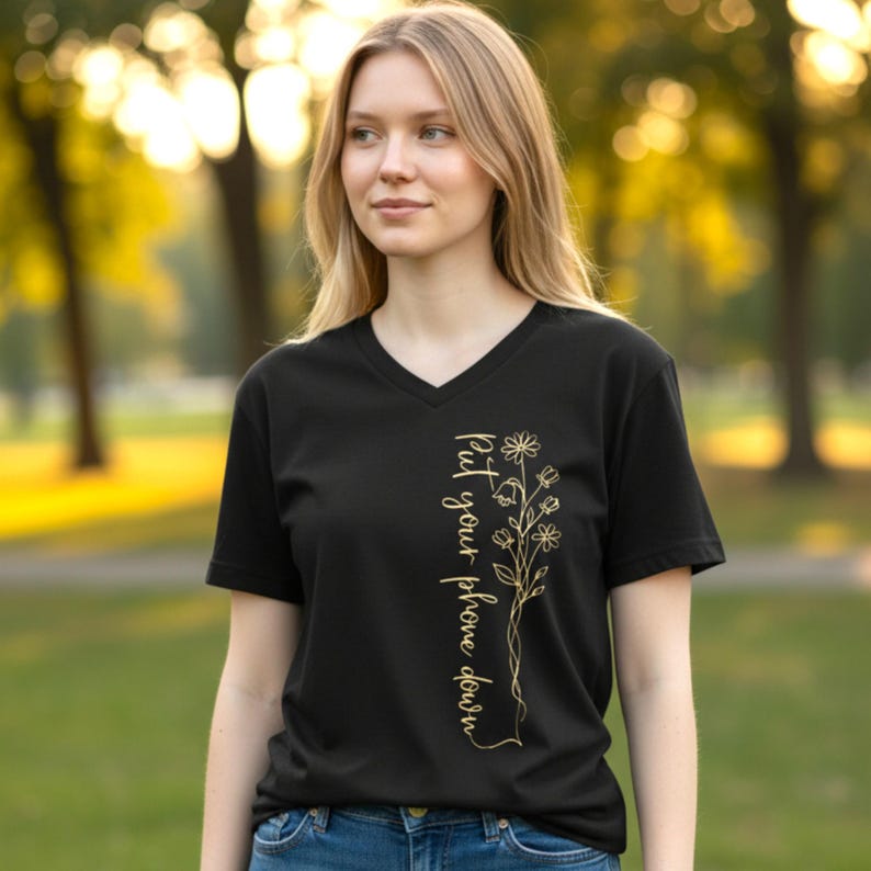 Puede incluir: Camiseta negra con cuello en V con dise&ntilde;o floral dorado y el texto "Put your phone down". El dise&ntilde;o presenta una disposici&oacute;n vertical de flores y hojas. La camiseta est&aacute; hecha de un material suave.