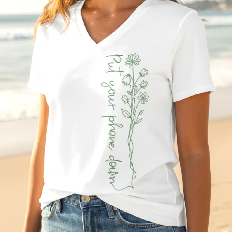 Puede incluir: Camiseta blanca con cuello en V con un dise&ntilde;o floral verde y el texto "Put your phone down" en escritura vertical. El dise&ntilde;o presenta flores y hojas delicadas. La camiseta est&aacute; hecha de un material suave.