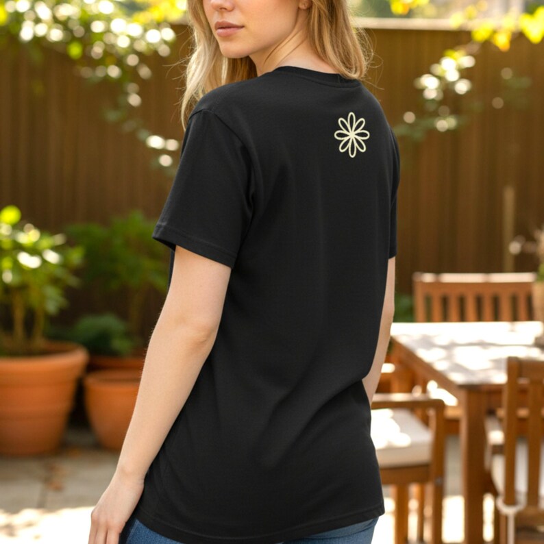 Puede incluir: Camiseta negra con un dise&ntilde;o de flor color crema en la espalda. La camiseta de manga corta est&aacute; hecha de un material suave. La persona est&aacute; de pie afuera cerca de una valla de madera y plantas en macetas.