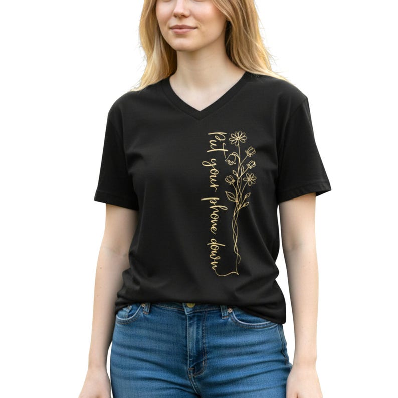 Puede incluir: Camiseta negra con cuello en V con texto dorado y dise&ntilde;o floral. El texto dice "Put your phone down" verticalmente en el lado izquierdo. El dise&ntilde;o floral tambi&eacute;n es dorado. La modelo lleva vaqueros azules.