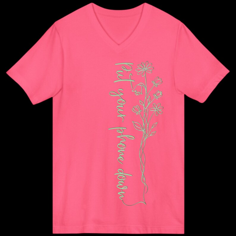 Puede incluir: Una camiseta rosa brillante con cuello en V con el texto "Put your phone down" en una fuente cursiva vertical. Un delicado dise&ntilde;o floral en color verde claro acompa&ntilde;a el texto. La camiseta es de color liso y tiene mangas cortas.