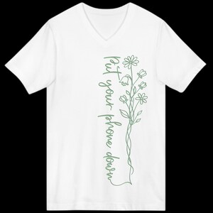 Puede incluir: Camiseta blanca con cuello en V con un dise&ntilde;o floral verde y el texto "put your phone down" en escritura vertical. El dise&ntilde;o presenta flores y hojas delicadas.