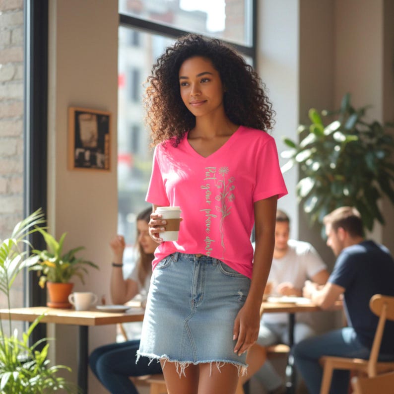 Puede incluir: Una mujer en un caf&eacute; con una camiseta rosa brillante con cuello en V y una falda vaquera. La camiseta tiene el texto "Put your phone down" y un dise&ntilde;o floral. Sostiene una taza de caf&eacute;.