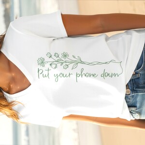 Puede incluir: Una camiseta blanca con cuello en V con un dise&ntilde;o floral verde y el texto "Put your phone down". El dise&ntilde;o presenta flores y hojas delicadas. La camiseta se combina con unos vaqueros azules.