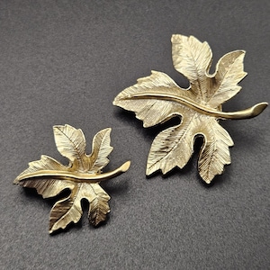 May include: Two gold-toned metal brooches shaped like maple leaves. Each brooch features a textured surface with fine etched lines mimicking leaf veins and a smooth, raised central stem that extends outward from the base of the leaf shape.