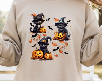 May include: Beige sweatshirt featuring a Halloween-themed graphic. The design includes three black cats wearing witch hats, sitting on carved pumpkins. Orange leaves and black bats are scattered around the cats, creating a festive autumn scene.