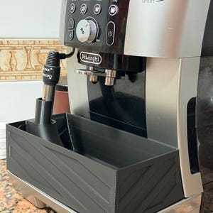 Puede incluir: Máquina de espresso De'Longhi Magnifica S Smart en negro y plateado. Cuenta con un espumador de leche y una bandeja negra en ángulo. El panel de control tiene opciones de café. Medidas en cm.
