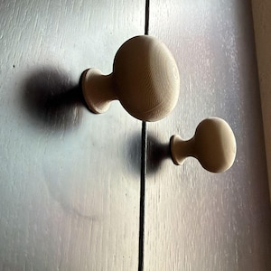 Könnte beinhalten: Zwei hölzerne Schrankknöpfe mit rundem Design sind an einer dunkelbraunen Holztür befestigt. Die Knöpfe sind hellbraun und haben eine glatte, kugelförmige Form. Die Tür hat eine vertikale Naht.