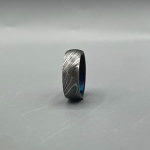 May include: A Damascus steel ring with a unique pattern. The ring has a dark gray exterior with a swirling, textured design. The interior of the ring is a vibrant blue color. This ring is a piece of jewelry.