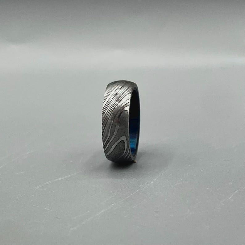 May include: A Damascus steel ring with a unique pattern. The ring has a dark gray exterior with a swirling, textured design. The interior of the ring is a vibrant blue color. This ring is a piece of jewelry.