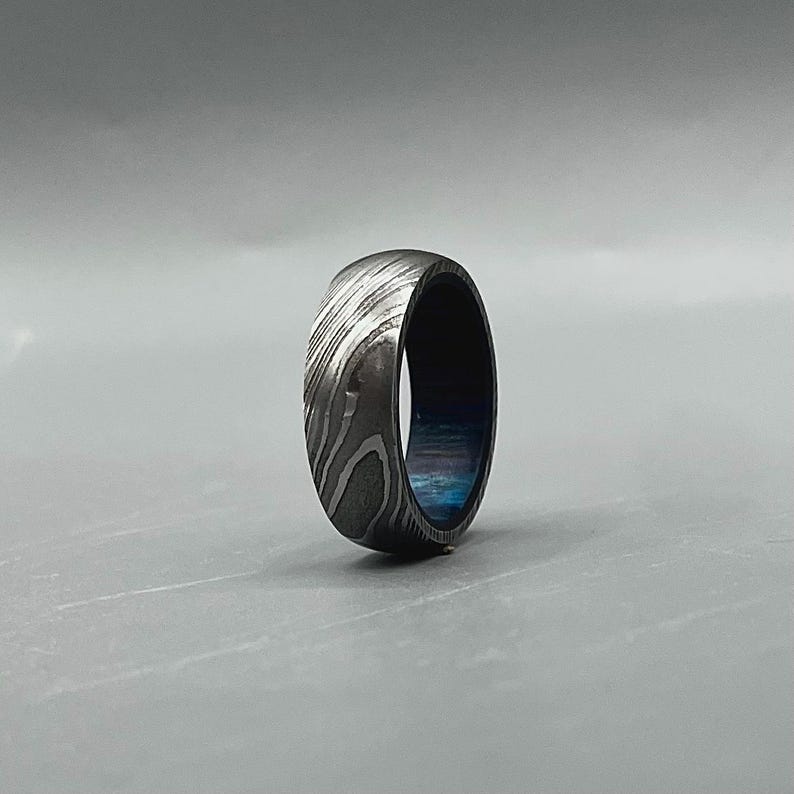 May include: A Damascus steel ring with a unique pattern of swirling light and dark metal. The ring has a dark blue interior and a rounded shape, showcasing a blend of textures and colors.