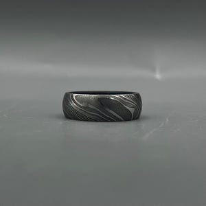 May include: A wide, dark gray ring with a unique, swirling pattern. The ring's surface has a textured appearance, with light and dark gray bands creating a wave-like design. The ring is set against a plain gray background.