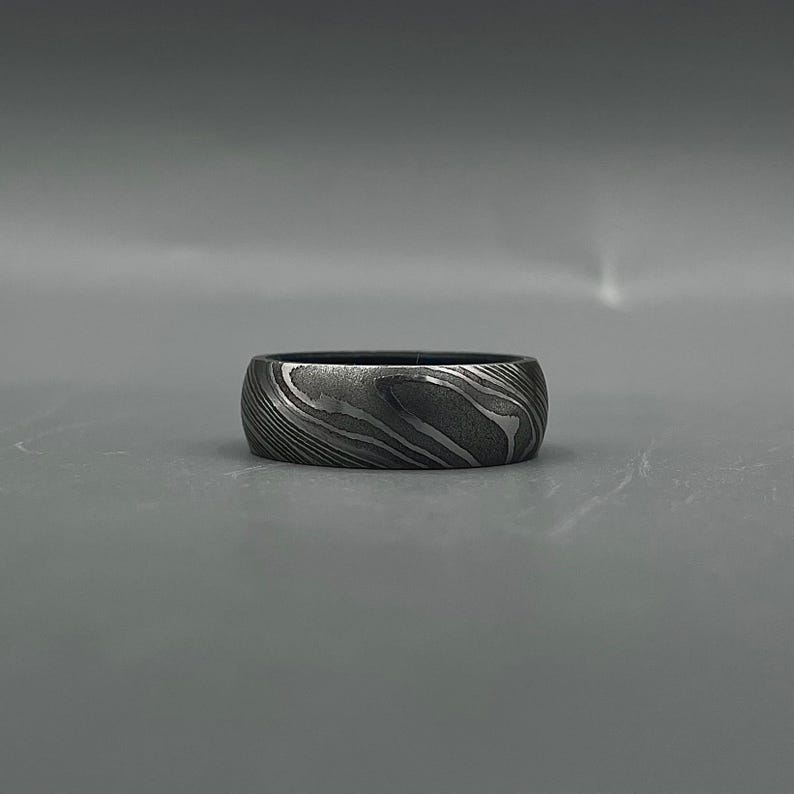 May include: A wide, dark gray ring with a unique, swirling pattern. The ring's surface has a textured appearance, with light and dark gray bands creating a wave-like design. The ring is set against a plain gray background.