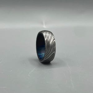May include: A Damascus steel ring with a swirling pattern and a blue interior. The ring is a band style, with a polished finish. The ring is shown on a gray background.