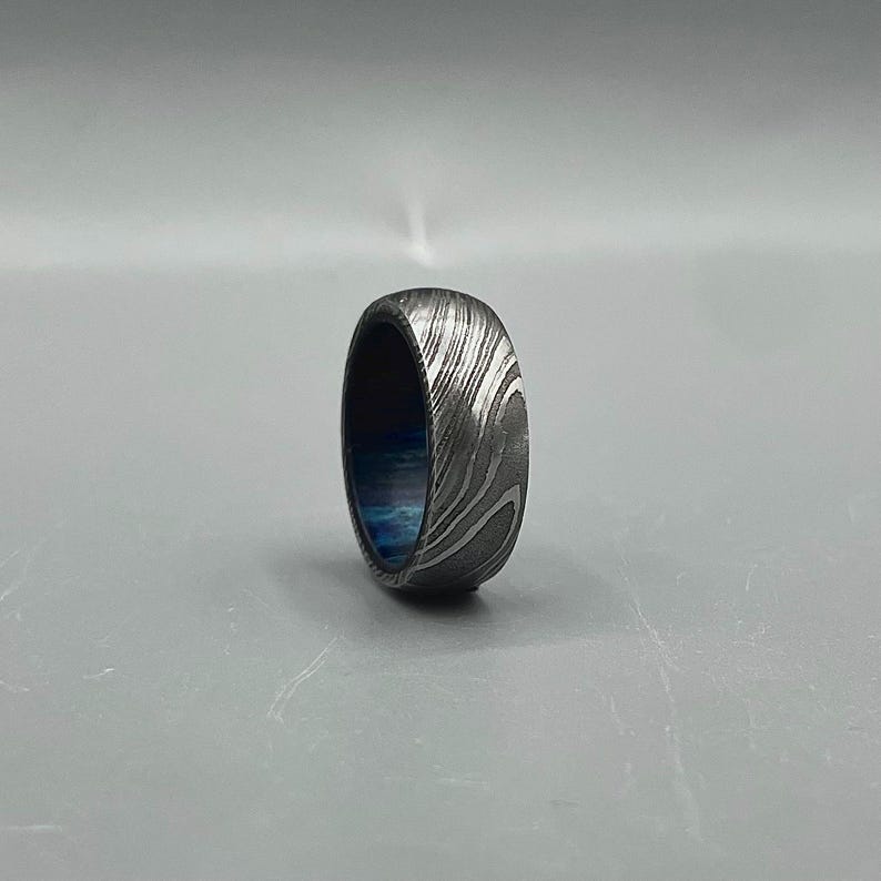 May include: A Damascus steel ring with a swirling pattern and a blue interior. The ring is a band style, with a polished finish. The ring is shown on a gray background.