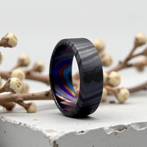 May include: A close-up of a unique ring with a dark, patterned exterior and a vibrant, multicolored interior. The ring's exterior features a dark gray and black striped design. The interior showcases a swirl of blue, purple, and orange hues.