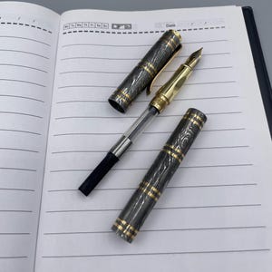 May include: A disassembled fountain pen with a black and gold patterned barrel and a gold nib. The pen is lying on a lined notebook page. The pen's cap is off, revealing the ink cartridge and nib.