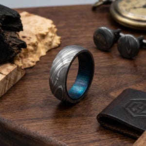 May include: A Damascus steel ring with a unique pattern and a blue interior. The ring is displayed on a wooden surface, alongside a pair of matching cufflinks and a leather wallet. The ring's design features a swirling, layered effect.