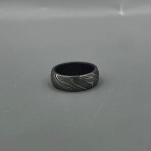 May include: A patterned metal ring with a dark interior. The ring has a swirling, wave-like design in shades of gray and silver. The ring is a band style, with a smooth, polished finish.