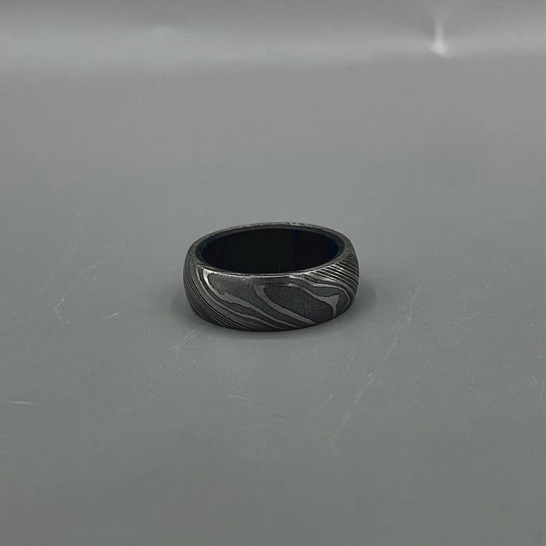 May include: A patterned metal ring with a dark interior. The ring has a swirling, wave-like design in shades of gray and silver. The ring is a band style, with a smooth, polished finish.