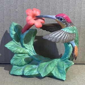 May include: A ceramic hummingbird figurine with a red head, green body, and brown wings. The bird is perched near a coral-colored flower and green leaves. The figurine is a decorative item.