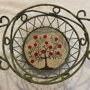 May include: A round wire basket with a distressed green finish and decorative scroll handles. The base features a circular ceramic insert with a crackled cream background and a painted tree illustration with green leaves and bright red fruit.