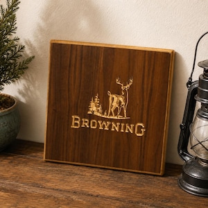 May include: A square dark brown wooden plaque featuring a laser-engraved design of a deer standing next to a pine tree. The word BROWNING is engraved in capital letters below the deer. The wood grain is visible across the flat surface.