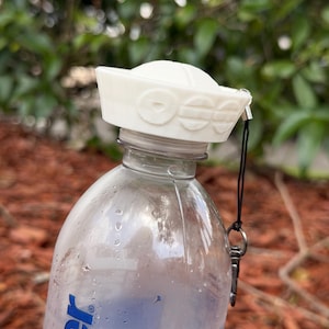 Peut inclure: Un bouchon de bouteille en forme de chapeau de marin blanc avec une calotte arrondie et un bord orné de détails circulaires en relief. Le bouchon est fixé à une bouteille d'eau en plastique transparent par un cordon noir et un clip.