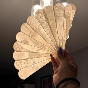 May include: A translucent, off-white folding fan featuring intricate, light-etched portraits of individuals across its multiple blades. The fan is held open, displaying detailed line work and the Japanese text 花様年華 vertically on the rightmost blade.
