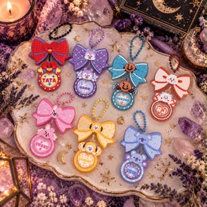 May include: A collection of eight colorful character keychains, each featuring a large bow attached to a circular charm. The characters include Tata, Mang, Shooky, RJ, Cooky, Chimmy, and Koya, each with a matching ball chain for hanging.